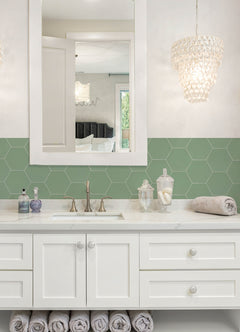 Modern kitchen featuring a white countertop, wooden stools with round seats and black footrests, a green backsplash made of WallPops Hexagon Peel & Stick Luxury Wall Tiles, light wood flooring, and a white backsplash. A blue pot and plants decorate the stove and counter. The space is illuminated by bright natural light from two windows.