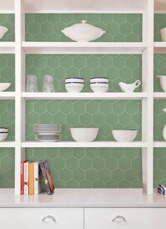 Modern kitchen featuring a white countertop, wooden stools with round seats and black footrests, a green backsplash made of WallPops Hexagon Peel & Stick Luxury Wall Tiles, light wood flooring, and a white backsplash. A blue pot and plants decorate the stove and counter. The space is illuminated by bright natural light from two windows.