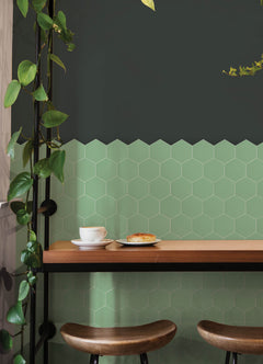 Modern kitchen featuring a white countertop, wooden stools with round seats and black footrests, a green backsplash made of WallPops Hexagon Peel & Stick Luxury Wall Tiles, light wood flooring, and a white backsplash. A blue pot and plants decorate the stove and counter. The space is illuminated by bright natural light from two windows.