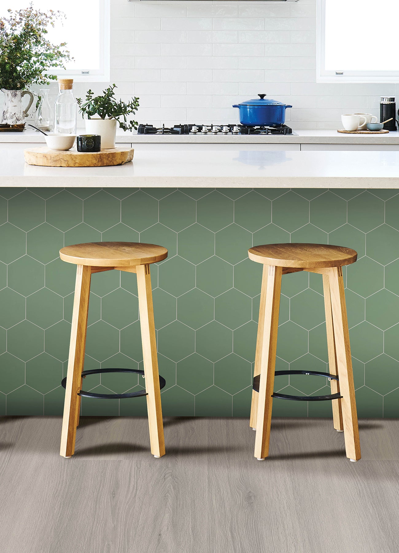 Modern kitchen featuring a white countertop, wooden stools with round seats and black footrests, a green backsplash made of WallPops Hexagon Peel & Stick Luxury Wall Tiles, light wood flooring, and a white backsplash. A blue pot and plants decorate the stove and counter. The space is illuminated by bright natural light from two windows.