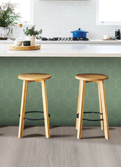 Modern kitchen featuring a white countertop, wooden stools with round seats and black footrests, a green backsplash made of WallPops Hexagon Peel & Stick Luxury Wall Tiles, light wood flooring, and a white backsplash. A blue pot and plants decorate the stove and counter. The space is illuminated by bright natural light from two windows.