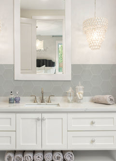 Modern kitchen featuring a white countertop, wooden stools with round seats and black footrests, a green backsplash made of WallPops Hexagon Peel & Stick Luxury Wall Tiles, light wood flooring, and a white backsplash. A blue pot and plants decorate the stove and counter. The space is illuminated by bright natural light from two windows.