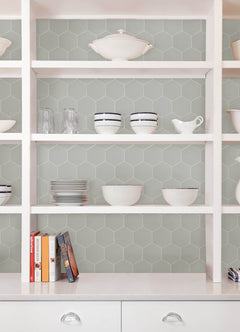 Modern kitchen featuring a white countertop, wooden stools with round seats and black footrests, a green backsplash made of WallPops Hexagon Peel & Stick Luxury Wall Tiles, light wood flooring, and a white backsplash. A blue pot and plants decorate the stove and counter. The space is illuminated by bright natural light from two windows.