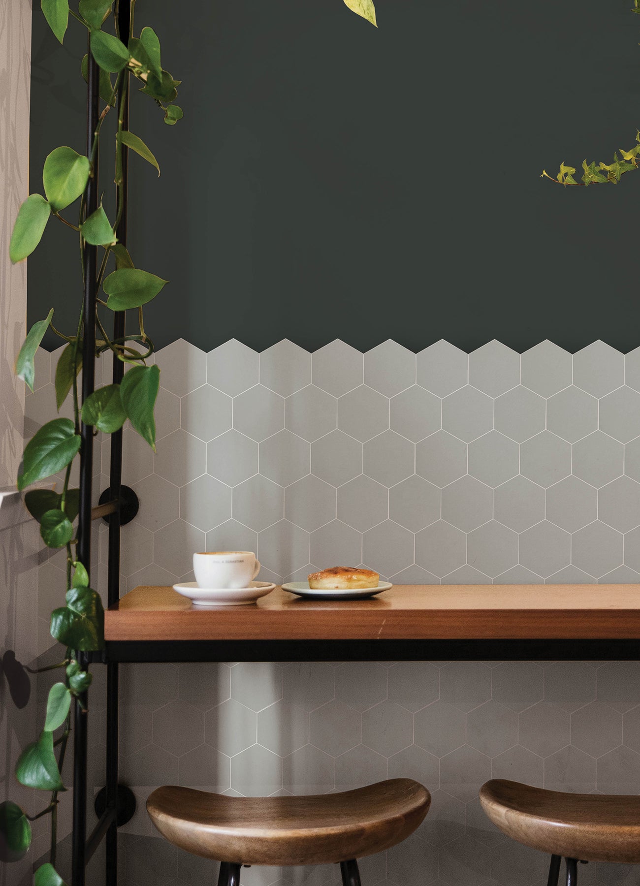 Modern kitchen featuring a white countertop, wooden stools with round seats and black footrests, a green backsplash made of WallPops Hexagon Peel & Stick Luxury Wall Tiles, light wood flooring, and a white backsplash. A blue pot and plants decorate the stove and counter. The space is illuminated by bright natural light from two windows.