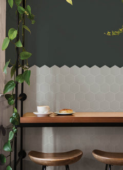 Modern kitchen featuring a white countertop, wooden stools with round seats and black footrests, a green backsplash made of WallPops Hexagon Peel & Stick Luxury Wall Tiles, light wood flooring, and a white backsplash. A blue pot and plants decorate the stove and counter. The space is illuminated by bright natural light from two windows.
