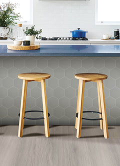 Modern kitchen featuring a white countertop, wooden stools with round seats and black footrests, a green backsplash made of WallPops Hexagon Peel & Stick Luxury Wall Tiles, light wood flooring, and a white backsplash. A blue pot and plants decorate the stove and counter. The space is illuminated by bright natural light from two windows.