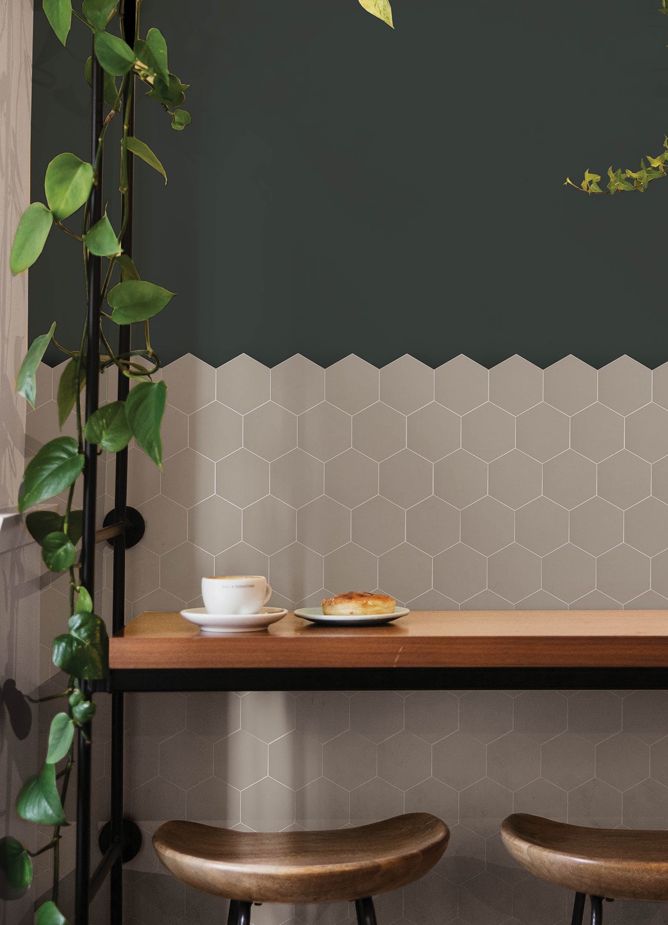 Modern kitchen featuring a white countertop, wooden stools with round seats and black footrests, a green backsplash made of WallPops Hexagon Peel & Stick Luxury Wall Tiles, light wood flooring, and a white backsplash. A blue pot and plants decorate the stove and counter. The space is illuminated by bright natural light from two windows.