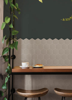 Modern kitchen featuring a white countertop, wooden stools with round seats and black footrests, a green backsplash made of WallPops Hexagon Peel & Stick Luxury Wall Tiles, light wood flooring, and a white backsplash. A blue pot and plants decorate the stove and counter. The space is illuminated by bright natural light from two windows.