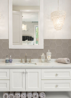 Modern kitchen featuring a white countertop, wooden stools with round seats and black footrests, a green backsplash made of WallPops Hexagon Peel & Stick Luxury Wall Tiles, light wood flooring, and a white backsplash. A blue pot and plants decorate the stove and counter. The space is illuminated by bright natural light from two windows.