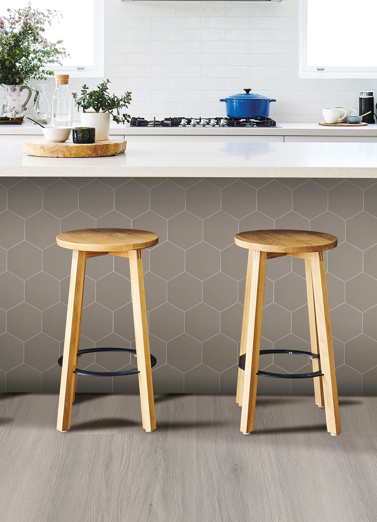 Modern kitchen featuring a white countertop, wooden stools with round seats and black footrests, a green backsplash made of WallPops Hexagon Peel & Stick Luxury Wall Tiles, light wood flooring, and a white backsplash. A blue pot and plants decorate the stove and counter. The space is illuminated by bright natural light from two windows.