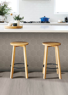 Modern kitchen featuring a white countertop, wooden stools with round seats and black footrests, a green backsplash made of WallPops Hexagon Peel & Stick Luxury Wall Tiles, light wood flooring, and a white backsplash. A blue pot and plants decorate the stove and counter. The space is illuminated by bright natural light from two windows.