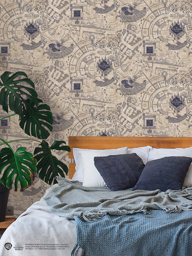 A cozy bedroom with a wooden bed, white and gray bedding, and dark gray pillows. A large green monstera plant sits by the bed. The wall has a detailed, vintage-style map wallpaper featuring castles and flying creatures in muted blue tones.