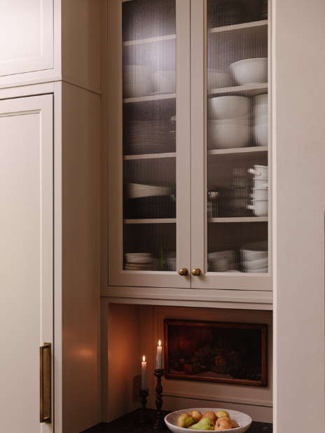 Tall cream kitchen cabinets with ribbed glass doors reveal neatly stacked dishware inside. Below, two lit candles in dark holders sit on a black counter. A bowl of apples and pears rests nearby. Warm, ambient lighting creates a cozy atmosphere.